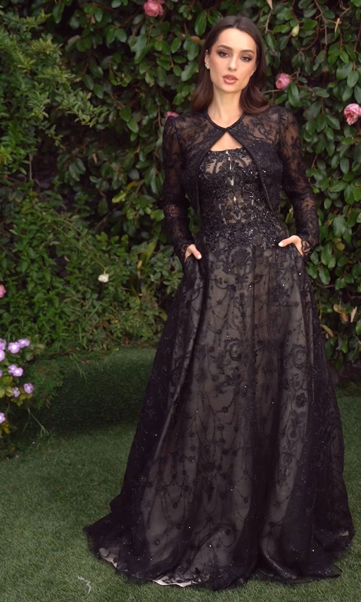 A woman stands on grass, wearing the Andrea & Leo MA104 Long Sleeve Long Formal Dress, featuring embroidered lace and intricate detailing, with green foliage and pink flowers in the background.